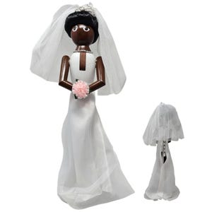 May include: A wooden bride figurine with a white dress, veil, and a pink flower bouquet. The figurine has dark brown arms and a black hairstyle. A smaller version of the figurine is also present, showcasing the same design.