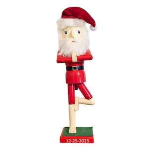 May include: A wooden Santa Claus figurine in a yoga pose. The figurine is dressed in a red suit with a black belt and a red hat. It has a white beard and is standing on a green base with the date 12-25-2025.