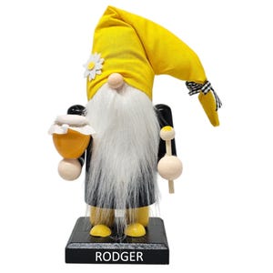 May include: A wooden nutcracker gnome with a yellow hat, black coat, and white beard. The gnome is holding a yellow drum and a wooden stick. The base of the nutcracker is black and has the word "RODGER" written on it.