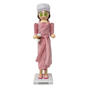May include: A wooden nutcracker figurine of a woman in a pink robe, eye mask, and a white towel on her head. She holds a coffee cup and wears pink sandals. The base reads "DONATA".