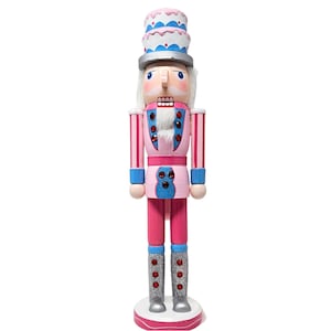 May include: A pink and blue wooden nutcracker with a white and blue cake hat. The nutcracker has a white beard and red gem accents.