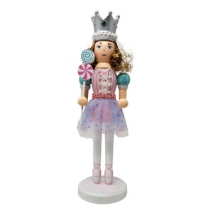 May include: A wooden nutcracker doll dressed as a ballerina with a pink and blue dress, a silver crown with a green jewel, and a pink and blue lollipop.