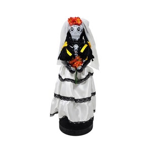 May include: A Day of the Dead bride doll with a white dress, black lace trim, and a veil. She is holding a bouquet of orange flowers and has a painted sugar skull face.