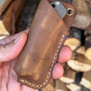 Peut inclure: Un étui de couteau en cuir marron avec un œillet en laiton et des coutures beiges. Un couteau gris et argenté est partiellement visible à l'intérieur. Le cuir présente un aspect vieilli.