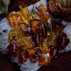 May include: A collection of amber-colored rune stones with white etched symbols. The stones vary in shape and shade, from light yellow to deep brown. They are arranged on a piece of birch bark, with a blurred background of leaves and a mushroom.