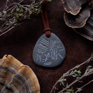 May include: A gray stone pendant with a carved design hangs from a brown leather cord. The pendant is surrounded by dried plants and mushrooms.