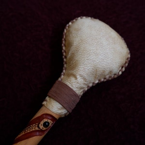 Puede incluir: Un sonajero con una cabeza redondeada, texturizada y de color beige claro unida a un mango de madera. El mango está envuelto con una banda marrón y decorado con un diseño de serpiente pintado en rojo y marrón con un detalle de ojo negro.