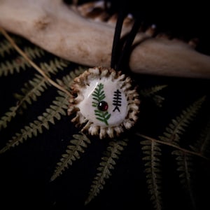 Op de afbeelding: Een handgemaakte hanger met een cirkelvormige geweibasis en een crèmekleurig midden. Het midden toont een groen varen- en zwart symboolontwerp, geaccentueerd door een kleine rode edelsteen. De hanger hangt aan een zwart koord, tegen een donkere achtergrond met varenbladeren.