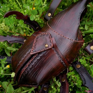 May include: A handcrafted, dark brown leather sheath with decorative stitching and brass accents. The sheath is designed to resemble a beetle and includes straps for carrying. The item is set against a backdrop of green foliage.
