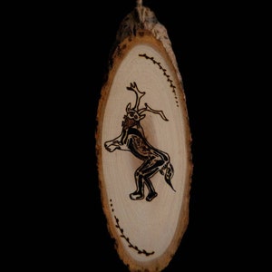 Pode incluir: Um ornamento de madeira com uma imagem queimada de uma criatura com chifres e um corpo humanoide. O ornamento tem forma oval e um acabamento de madeira natural.