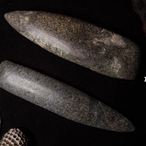 May include: Two gray stone tools with rounded ends, possibly pestles or grinding tools, are displayed against a dark background. One is labeled "1" and the other "2". The tools have a smooth, polished appearance.