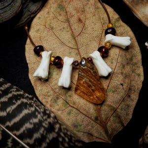 May include: A necklace featuring a central amber-colored pendant, four white bone-shaped beads, and several small dark brown spherical beads strung on a thin cord. The necklace is arranged on a large, dried, veined brown leaf.