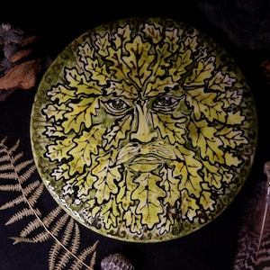 May include: A round, ceramic plate featuring a Green Man design. The face is surrounded by yellow-green leaves with black outlines. The plate has a green border and is set against a dark background with natural elements.