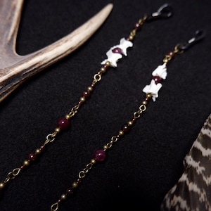 May include: A pair of long earrings featuring antique brass chains, small white bone segments, and deep red garnet-colored beads. Each earring is finished with a black metal hook at the top, creating a linear design with alternating metal and stone elements.