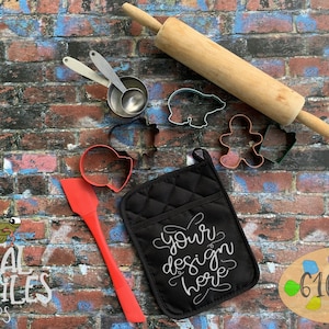 May include: A black oven mitt with the text "your design here" in white on a brick background.  There are various kitchen tools and cookie cutters scattered around the mitt, including a rolling pin, measuring cups, and spatulas.