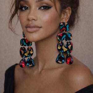 May include: Long, dangling earrings with a black base and colorful abstract shapes in red, teal, and gold. The earrings have a layered design with various geometric forms, creating a bold and artistic statement. The earrings are a fashion accessory.