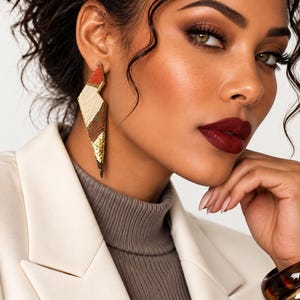 May include: A long, geometric, lightning-bolt-shaped earring featuring a textured, multi-colored surface with segments of cream, rust orange, dark brown, and gold metallic foil. The earring hangs vertically from the earlobe with a sharp, pointed tip.