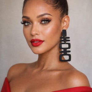 May include: A woman with dark hair styled in a bun, wearing bold black geometric earrings and a red off-the-shoulder top. Her makeup includes red lipstick and dramatic eye makeup. The background is a neutral color.