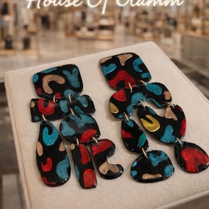May include: Black dangle earrings with abstract shapes in red, teal, and gold patterns. The earrings are connected with gold-colored metal. The text "House Of Glamm" is in the background.
