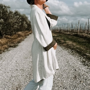 May include: A black wide-brimmed hat, a long-sleeved white cardigan with dark olive green cuffs, a black top, light-wash denim jeans with a raw hem, and black and white leopard print high-top sneakers worn on a gravel path.