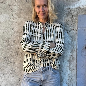Op de afbeelding: Een vrouw draagt een crèmekleurige blouse met een groen geometrisch patroon en lichtblauwe spijkerbroekshorts. De blouse heeft lange mouwen en een strikdetail in de hals. Ze staat voor een grijze, getextureerde muur.