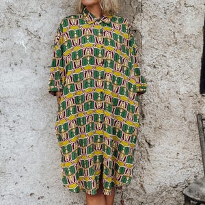 Puede incluir: Vestido con cuello y botones con un patrón repetido de formas verdes y amarillas sobre un fondo granate. El vestido llega a la rodilla y tiene mangas tres cuartos. La persona está descalza frente a una pared clara y texturizada.