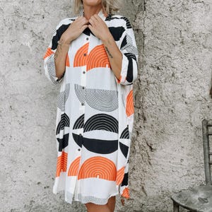 Puede incluir: Vestido camisero blanco con un llamativo estampado geométrico de semicírculos naranjas, negros y blancos. El vestido tiene mangas tres cuartos y llega hasta la pantorrilla. Una silla de madera desgastada está a la derecha.