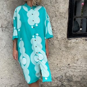 May include: A turquoise button-down shirt dress with a white abstract pattern. The dress has short sleeves and a knee-length hem. The model is standing barefoot against a textured stone wall.