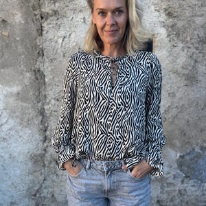 Pode incluir: Uma mulher usando uma blusa preta e branca com estampa de zebra, mangas compridas e decote em forma de fechadura. Ela também está usando jeans azul claro. A blusa tem um caimento solto e descontraído. O fundo é uma parede de pedra cinza texturizada.