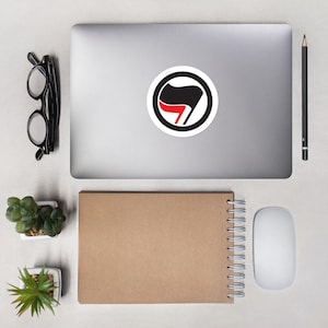 May include: A black and red anarchist symbol sticker on a silver laptop computer. The sticker is a circle with a black fist holding a red flag.