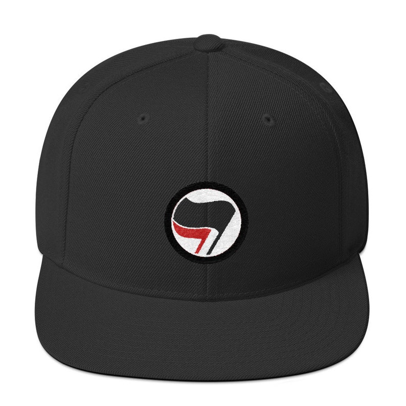 May include: Black and white flat brimmed baseball cap with a red and black circle logo featuring a stylized flag with a red stripe.