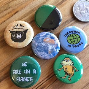 May include: Nine round button pins with various designs.  One pin features a green and black color scheme.  Another pin features a green globe on a blue background with the text "THE POWER IS YOURS".  A third pin features a cartoon character with a bushy beard and the text "WE ARE ON A PLANET".  A fourth pin features a cartoon bear wearing a ranger hat with the text "EAT THE RICH".  A fifth pin features a blue and white image of the Earth.  A sixth pin features a green background with a cartoon character with a bushy beard.