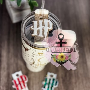 May include: A gold glitter tumbler with a white and gold glitter monogram charm that says "HP". The tumbler has a straw and a white lid. The tumbler is sitting on a wooden surface. The image also includes two other charms with the same monogram, one in red and white stripes and one in green and white stripes.