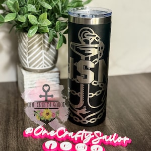 May include: Black insulated tumbler with a silver lid, featuring a USN anchor design. The tumbler has the text "USN" in a silver color. The image also includes a small potted plant and a pink sign that says "One Crafty Sailor".