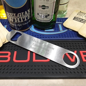 May include: A silver bottle opener with a personalized inscription that reads "Michael Johnson Groomsman June 25, 2020". The bottle opener is lying on a black and red bar mat.