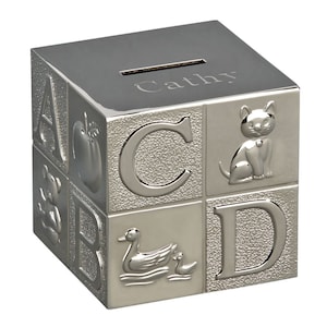 May include: A silver-colored, cube-shaped bank with embossed letters, a cat, a duck with a duckling, and an apple. The top of the bank has a slot for inserting money and the name "Cathy" engraved on it.