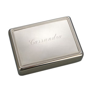 May include: A rectangular silver-colored box with a beaded border and the name "Cassandra" engraved on the lid. The box has a shiny, reflective surface and a dark gray base.