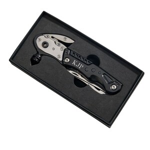 May include: A black multi-tool with a silver blade and a hammer head. The tool is engraved with the initials "KJP" and is in a black box.