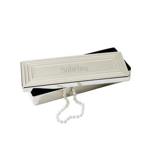 May include: A silver rectangular jewelry box with the name "Sabrina" engraved on the lid. The box has a decorative border and is lined with black velvet. A strand of white pearls hangs from the box.