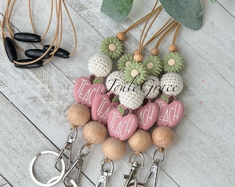 May include: A collection of teacher lanyards with various designs. The lanyards feature wooden beads, silver clasps, and decorative elements like pink apple-shaped charms with the word "TEACH", green daisy-shaped beads, and white crocheted beads.