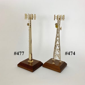 May include: Two gold metal model cell phone towers on wooden bases. The tower on the left is tall and thin with multiple antennas. The tower on the right is shorter and wider with a triangular shape. The bases are dark brown wood. The towers are labeled #477 and #474.