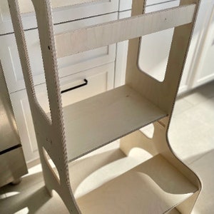 Peut inclure: Un tabouret d'apprentissage en bois avec deux étagères et une découpe sur le côté. L'étagère supérieure porte le nom "August" gravé.
