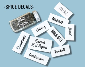 May include: White labels with black text for various spices, including black pepper, nutmeg, cinnamon, paprika, oregano, garlic salt, basil, crushed red pepper, cardamom, and sea salt. -SPICE DECALS- is written at the top of the image.