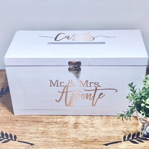 May include: White wooden box with a slot on the top for cards. The box has a gold inscription that reads "Cards" and "Mr. & Mrs. Aponte".