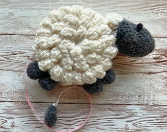 May include: A white crocheted sheep with gray accents, including a face, legs, and tail. The sheep is attached to a pink and white measuring tape.