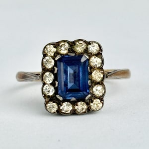 May include: A vintage ring featuring a rectangular, deep blue gemstone surrounded by a halo of smaller, clear stones. The ring has a gold-toned band and a dark, ornate setting around the central stone, creating a classic, elegant look.