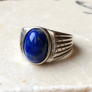 Puede incluir: Un anillo de plata con una piedra ovalada de lapislázuli azul. El anillo tiene una banda texturizada.