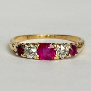 Może przedstawiać: Złoty pierścionek z centralnym owalnym rubinem, otoczonym dwoma okrągłymi, przezroczystymi diamentami i dwoma mniejszymi rubinami. Obrączka ma ozdobny, grawerowany wzór. Pierścionek jest na jasnym tle.