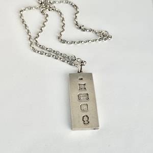 May include: A silver-toned pendant necklace with a rectangular pendant and a chain. The pendant has several engraved hallmarks. The chain is a simple, linked design. The necklace is displayed on a white surface.