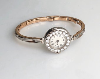 May include: A vintage wristwatch with a white face and black Roman numeral hour markers. The watch has a round, diamond-encrusted bezel and a two-tone gold band.